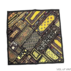 Op de afbeelding: Een vierkante patchworktextiel met een zwarte rand. Het ontwerp bevat verschillende geometrische patronen in geel, zwart, bruin en crème. De textiel heeft een vintage uitstraling.