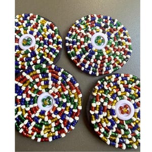 May include: Four circular beaded coasters. Each coaster has a spiral pattern of red, white, blue, green, and gold beads. A white button with a floral design is in the centre of each coaster. The coasters are on a dark grey surface.
