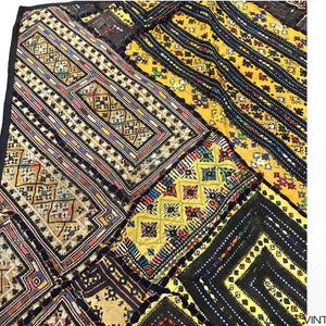 Op de afbeelding: Een patchwork textiel met diverse geometrische patronen en kleuren. Het ontwerp omvat rechthoekige en ruitvormige vormen in tinten geel, zwart, bruin en beige. Het textiel heeft een zwarte rand en lijkt vintage te zijn.