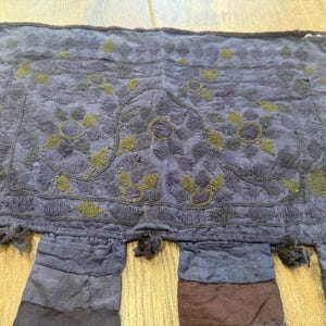 Könnte beinhalten: Ein rechteckiges, indigofarbenes Textil mit aufwendiger Stickerei. Das Design zeigt Blumen- und Blättermuster in Olivgrün und Lila. Der untere Rand hat mehrere hängende Stoffstreifen und Quasten.