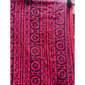May include: A red textile featuring a repeating pattern of dark blue circles, diamonds, and vertical lines. The fabric is likely cotton or a similar material, and the design is symmetrical. The textile is hanging from a black clip.