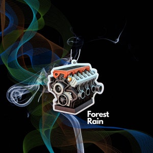 Könnte beinhalten: Ein weißer Autolufterfrischer in Form eines detaillierten Motors mit orangefarbenen Akzenten. Der Text "Forest Rain" ist unter dem Motor aufgedruckt.