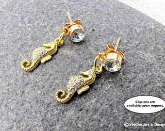 May include: A pair of gold-colored seahorse earrings with clear crystal accents. The seahorses are embellished with small, sparkling crystals. The earrings have a post-back style with a large, clear crystal. Text on the image: "Clip-ons are available upon request."