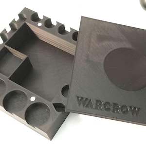 May include: A black organizer with a lid, featuring compartments and circular cutouts. The lid has a circular indentation and the word "WARGROW" embossed on it. Ideal for storing small items.