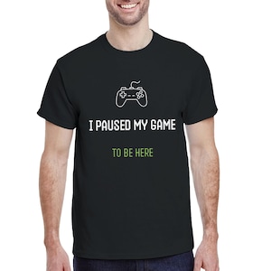 May include: Black t-shirt featuring a white game controller graphic and the text "I PAUSED MY GAME TO BE HERE" in white and green. The shirt is made of a soft material.