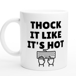 May include: White ceramic mug with black text that reads "THOCK IT LIKE IT'S HOT" and a black and white illustration of a keyboard with two hands typing.