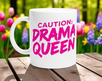 Op de afbeelding: Witte keramische mok met de tekst "CAUTION: DRAMA QUEEN" in felroze letters. De mok heeft een gebogen oor en staat op een houten oppervlak met een vage bloemenachtergrond.