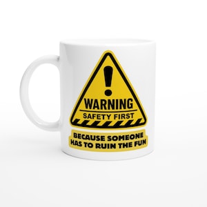 May include: A white ceramic mug featuring a yellow triangular warning sign graphic with a black exclamation point. The text reads WARNING SAFETY FIRST and BECAUSE SOMEONE HAS TO RUIN THE FUN in bold black lettering with a striped border detail.