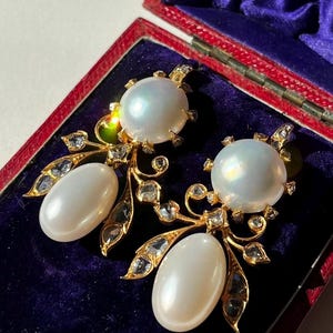 以下が含まれることがあります： 大きな白い真珠と小さな透明な宝石をあしらった、金色のイヤリングのペア。イヤリングは、上部に大きな丸い真珠、下部に楕円形の真珠が付いた、華やかなフローラルデザインです。赤いジュエリーボックスに展示されています。
