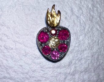May include: A heart-shaped pendant with a gold flame at the top. The pendant is silver with a border of small stones. The heart is filled with pink and yellow gemstones. Jewelry.