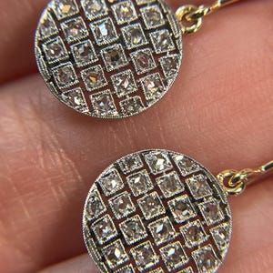 May include: Close-up of a pair of gold earrings. Each earring has a circular design with a grid pattern of square-cut, clear gemstones. The earrings are suspended from delicate gold wires, showcasing a classic jewellery style.