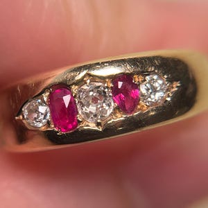 May include: A gold band ring featuring a row of five alternating gemstones. The setting includes two round clear stones, two oval-shaped vibrant red rubies, and one central round clear stone, all securely mounted within the polished gold metal band.