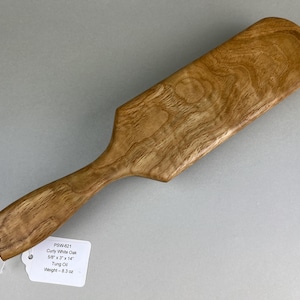 May include: A handcrafted wooden spatula made of curly white oak. The spatula is 14 inches long and 3 inches wide, with a handle and a paddle-like head. The wood grain is visible, and the spatula has a hole in the handle for hanging.