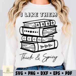 Può includere: Una felpa bianca con un design grafico nero di una pila di libri. Il testo "I like them thick & spicy" è stampato sotto i libri.