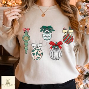 Könnte beinhalten: Ein cremefarbener Pullover mit einem Design aus sechs Weihnachtskugeln. Die Kugeln sind in verschiedenen Farben und Mustern, darunter Rot, Grün, Weiß und Silber. Die Kugeln sind alle in verschiedenen Formen und Größen.