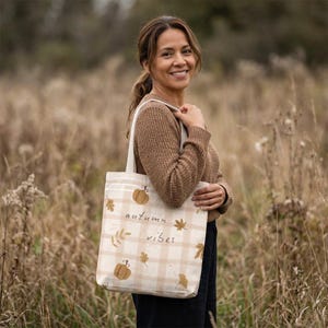Könnte beinhalten: Eine beigeweiße karierte Tragetasche mit Illustrationen von orangefarbenen Kürbissen und braunen Herbstblättern. Der Text "autumn vibes" ist in einer einfachen dunklen Schrift auf der Vorderseite der Tasche gedruckt, die zwei Schulterriemen hat.