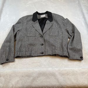 May include: A gray blazer with a black collar and buttons. The blazer has two front pockets and long sleeves with black buttons at the cuffs. The jacket is made of a textured fabric.