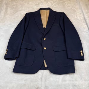 Puede incluir: Un blazer azul marino con botones dorados y un interior beige. El blazer tiene dos bolsillos delanteros y tres botones en cada puño. La chaqueta está colocada sobre una superficie blanca.