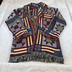 May include: A woven open-front jacket with a patchwork design featuring red, white, and blue patterns. The jacket has a fringed hem and sleeve edges. The design includes stars, stripes, and geometric shapes, creating a patriotic theme.