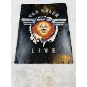 Può includere: Un programma di concerti in bianco e nero con la scritta "VAN HALEN LIVE". La copertina presenta un leone ruggente con gli occhiali, che sfonda un emblema bianco e argento. Il leone ha una pelliccia arancione e marrone.