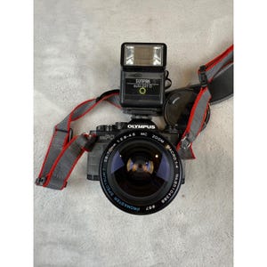 Puede incluir: Cámara Olympus negra con flash Sunpak y correa gris y roja. La lente de la cámara dice "28-70mm 1:2.8-4.5 MC ZOOM MACRO 1:4". El flash dice "SUNPAK auto 221 D".