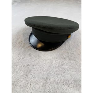 Puede incluir: Un sombrero de estilo militar verde oscuro con una visera negra brillante. El sombrero tiene una parte superior redondeada y un botón dorado en el lateral. El sombrero está sobre una superficie gris claro.