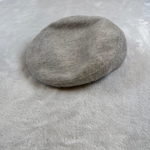 Pode incluir: Uma boina cinza claro, feita de um material semelhante à lã. O chapéu tem uma coroa arredondada e uma aba pequena e rígida. A boina é de cor sólida e parece ser feita de um tecido macio e texturizado.