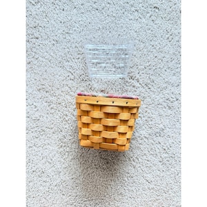Puede incluir: Una cesta de madera trenzada de color marrón claro con un borde plano que presenta una cenefa de tela a cuadros rojos, verdes y blancos. Sobre la cesta hay un forro de plástico transparente rectangular con un patrón de rejilla.
