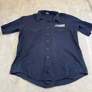 Puede incluir: Una camisa de manga corta en color azul marino oscuro con cuello, dos bolsillos en el pecho con solapas abotonadas y un parche con el logotipo de United States Postal Service en el pecho izquierdo. Tiene una fila central de botones.