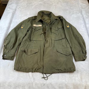 May include: An olive green military jacket with multiple pockets, a drawstring waist, and a collared neckline. The jacket has patches on the sleeves and a name tag that reads "BURT". The jacket is made of a durable fabric.
