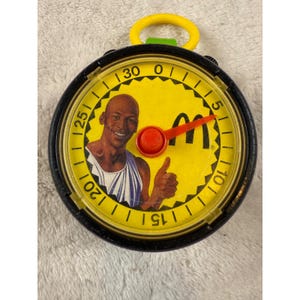 Può includere: Un cronometro vintage di McDonald's con un quadrante giallo raffigurante un uomo sorridente che fa il pollice in su. Il cronometro ha una lunetta nera, un pulsante rosso e un anello giallo. Il logo McDonald's è visibile sul quadrante.