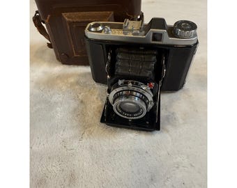 May include: A vintage black and silver camera with a leather case. The camera has a retractable lens and various dials and settings. The leather case is brown and has a flap closure. The camera is a classic photography item.