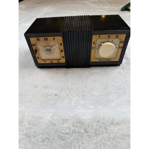 以下が含まれることがあります： ヴィンテージの黒とタンのFirestoneクロックラジオ。時計の文字盤はタン色で、黒い数字と針があります。ラジオの文字盤はタン色で、大きなクリーム色のノブとFirestoneのロゴが付いています。時計はユニークなアコーディオン風のデザインです。