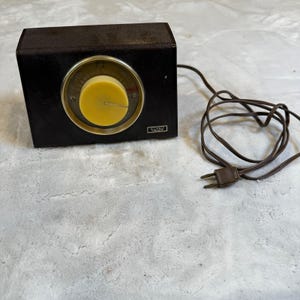 May include: A vintage brown box with a yellow dial and a power cord. The dial has a white needle and a circular design. The box has a small rectangular label that reads "ALLIANCE".