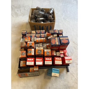 May include: A collection of vintage vacuum tubes and their original packaging. The boxes are in various sizes and colors, with prominent branding from GE and RCA. A cardboard box filled with tubes sits above the display.