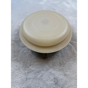 Puede incluir: Un recipiente de plástico redondo de color beige claro con tapa. La tapa tiene un centro ligeramente elevado con un pequeño diseño circular. El recipiente parece estar hecho de un material plástico mate duradero, adecuado para el almacenamiento de alimentos.