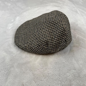 May include: A tweed newsboy cap in a houndstooth pattern. The cap features a classic, rounded shape with a small, stiff brim. The fabric is a mix of black, brown, and beige, creating a textured appearance. The cap is likely made of wool or a wool blend.