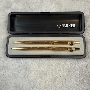 Könnte beinhalten: Ein goldfarbenes Parker Stift- und Bleistiftset in einem schwarzen Etui. Das Etui ist geöffnet und zeigt die Schreibgeräte. Das Parker-Logo ist weiß auf der Innenseite des Etuis aufgedruckt.