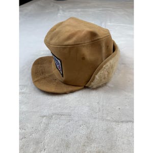 May include: A tan-colored winter hat with a curved brim and ear flaps. The hat features a patch with a design in red, white, and blue. The ear flaps are lined with a cream-colored, fuzzy material.