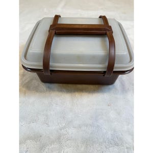 Può includere: Un portapranzo quadrato marrone e beige con un manico in pelle marrone. Il portapranzo ha una base marrone e un coperchio beige. Il manico è attaccato al coperchio con cinghie in pelle marrone. Il portapranzo è su una superficie bianca.