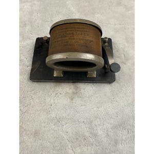 May include: An antique magnetizer with a cylindrical brown body and a silver-colored rim. The device is mounted on a black rectangular base with a handle and a small circular knob. Text is visible on the side of the cylinder.