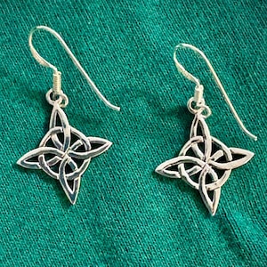 May include: A pair of silver-colored earrings with a Celtic knot design. The earrings have a hook closure and a four-pointed knot pendant. The earrings are displayed on a green fabric surface.