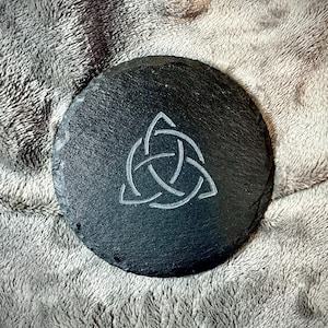 May include: A round, black slate coaster with a white Celtic knot design etched into the surface. The coaster is approximately 4 inches in diameter and has a rough, textured appearance. The coaster is a decorative item.
