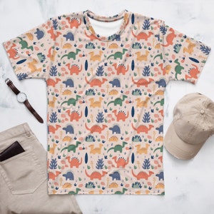 Può includere: Una maglietta rosa chiaro con un motivo ripetuto di dinosauri colorati e piante verdi. I dinosauri sono arancioni, gialli, verdi e blu.