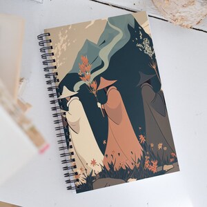 May include: A spiral bound notebook with a colorful illustration of three figures in hooded cloaks walking through a misty landscape. The figures are silhouetted against a dark blue background with a mountain range in the distance. The notebook cover features a whimsical and stylized design.