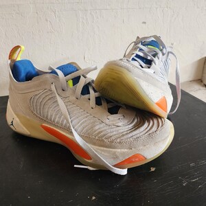 Puede incluir: Par de zapatillas deportivas grises y blancas con detalles en naranja y azul. Las zapatillas tienen cordones blancos y un logotipo Jumpman visible. Las suelas son de color marr&oacute;n claro.