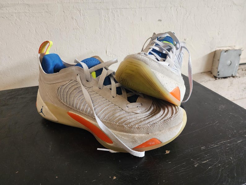 Puede incluir: Par de zapatillas deportivas grises y blancas con detalles en naranja y azul. Las zapatillas tienen cordones blancos y un logotipo Jumpman visible. Las suelas son de color marr&oacute;n claro.