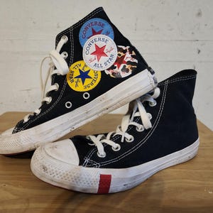 May include: A pair of black high-top sneakers with white laces and white rubber soles. The side of one sneaker features three circular patches: a blue one with a red star, a white one with a red star, and a yellow one with a blue star.