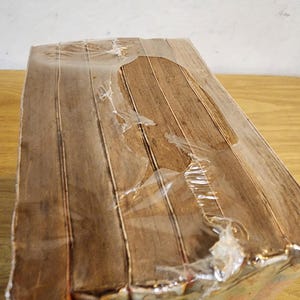 May include: A rectangular, wrapped package with a wood-grain pattern. The package is sealed in clear plastic, with some tears in the wrapping. The wood-grain pattern is a light brown color.