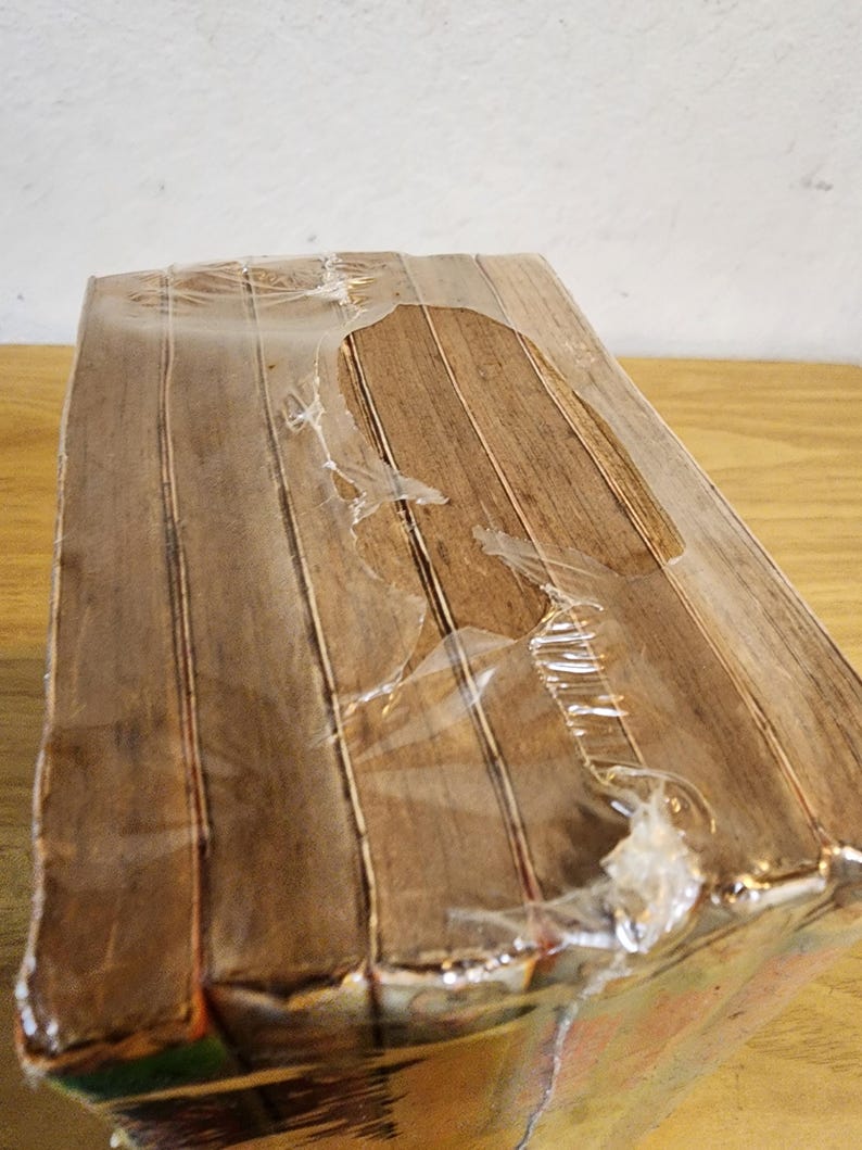 May include: A rectangular, wrapped package with a wood-grain pattern. The package is sealed in clear plastic, with some tears in the wrapping. The wood-grain pattern is a light brown color.